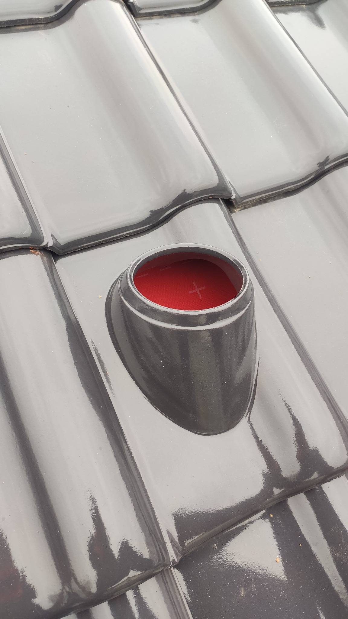 Błyszcząca, grafitowa dachówka ceramiczna z okrągłym kominkiem wentylacyjnym z czerwonym filtrem wewnątrz.