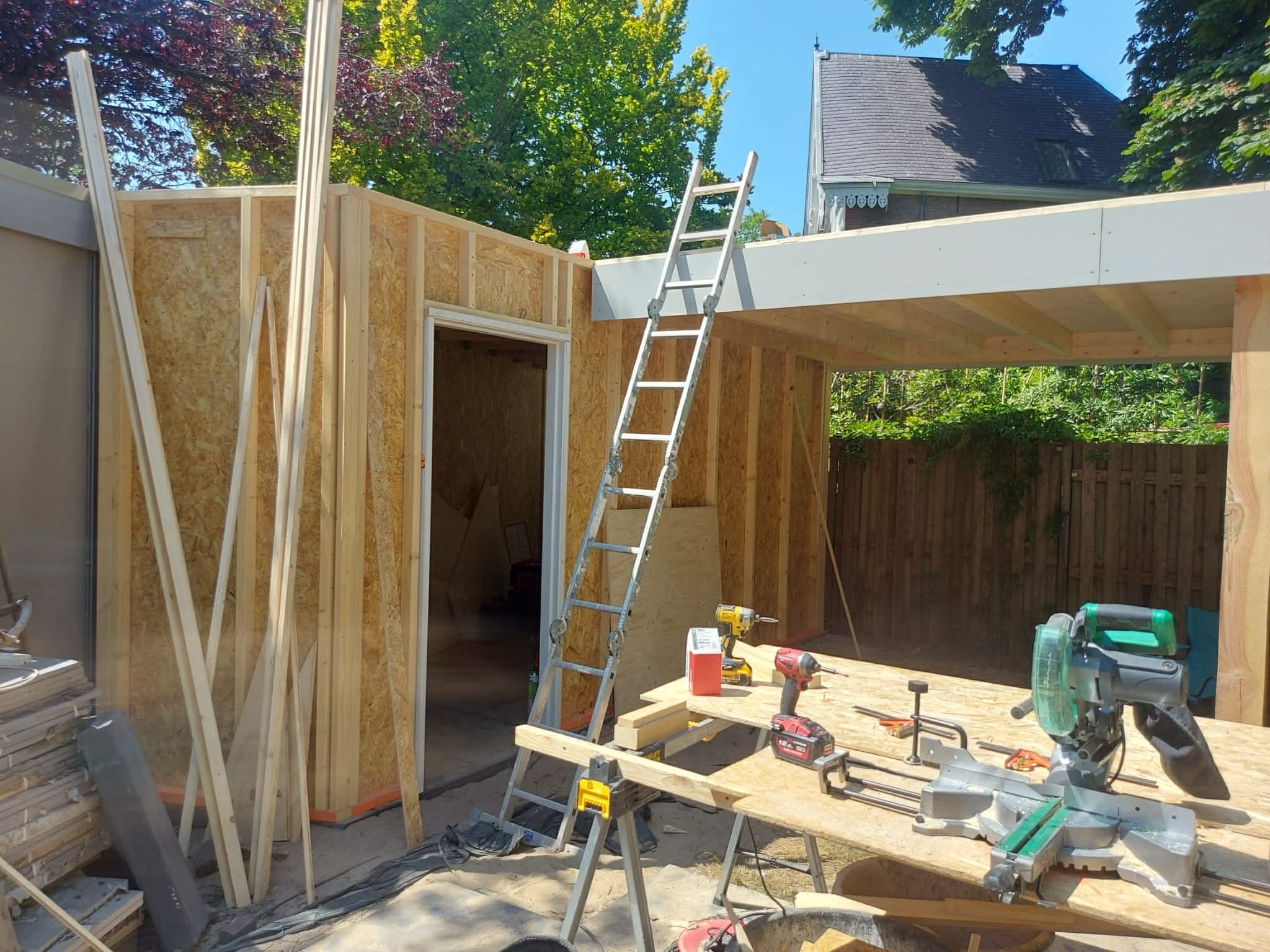 Budowa szkieletowej konstrukcji budynku z drewna, widoczne narzędzia i drabina. Prace w toku, jasne oświetlenie dzienne, zielone drzewa w tle.