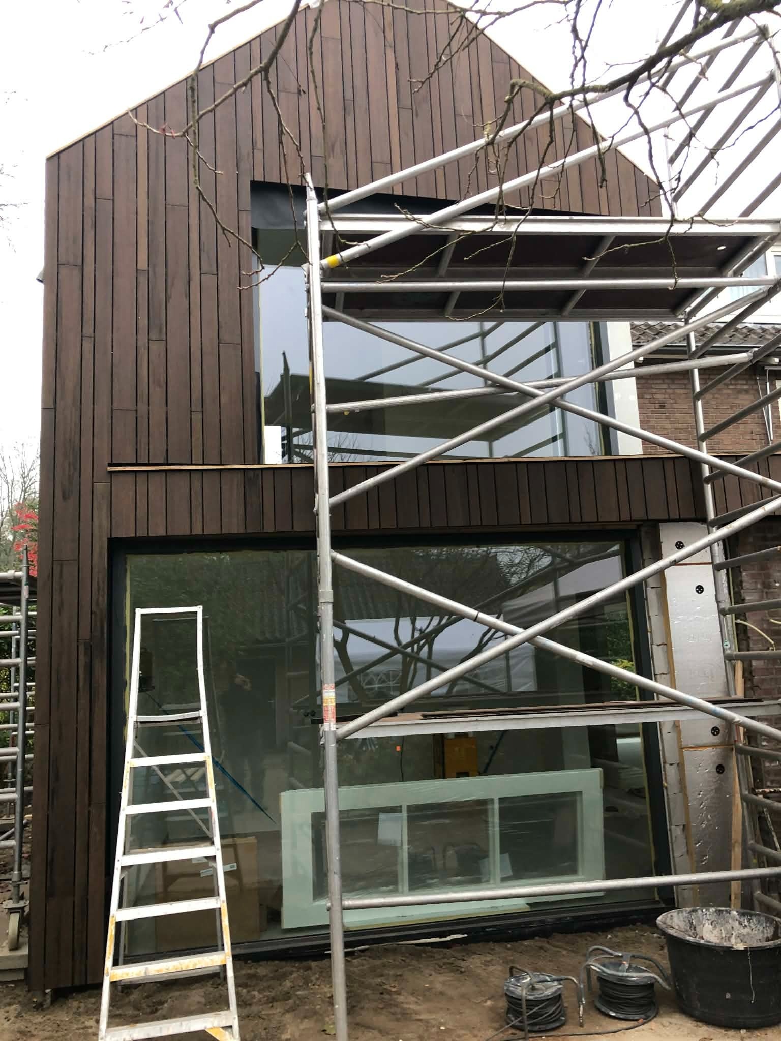 Nowoczesny dom z elewacją z drewnianych paneli w trakcie montażu. Widoczne rusztowania, okna i drabina. Prace wykończeniowe na zewnątrz budynku.