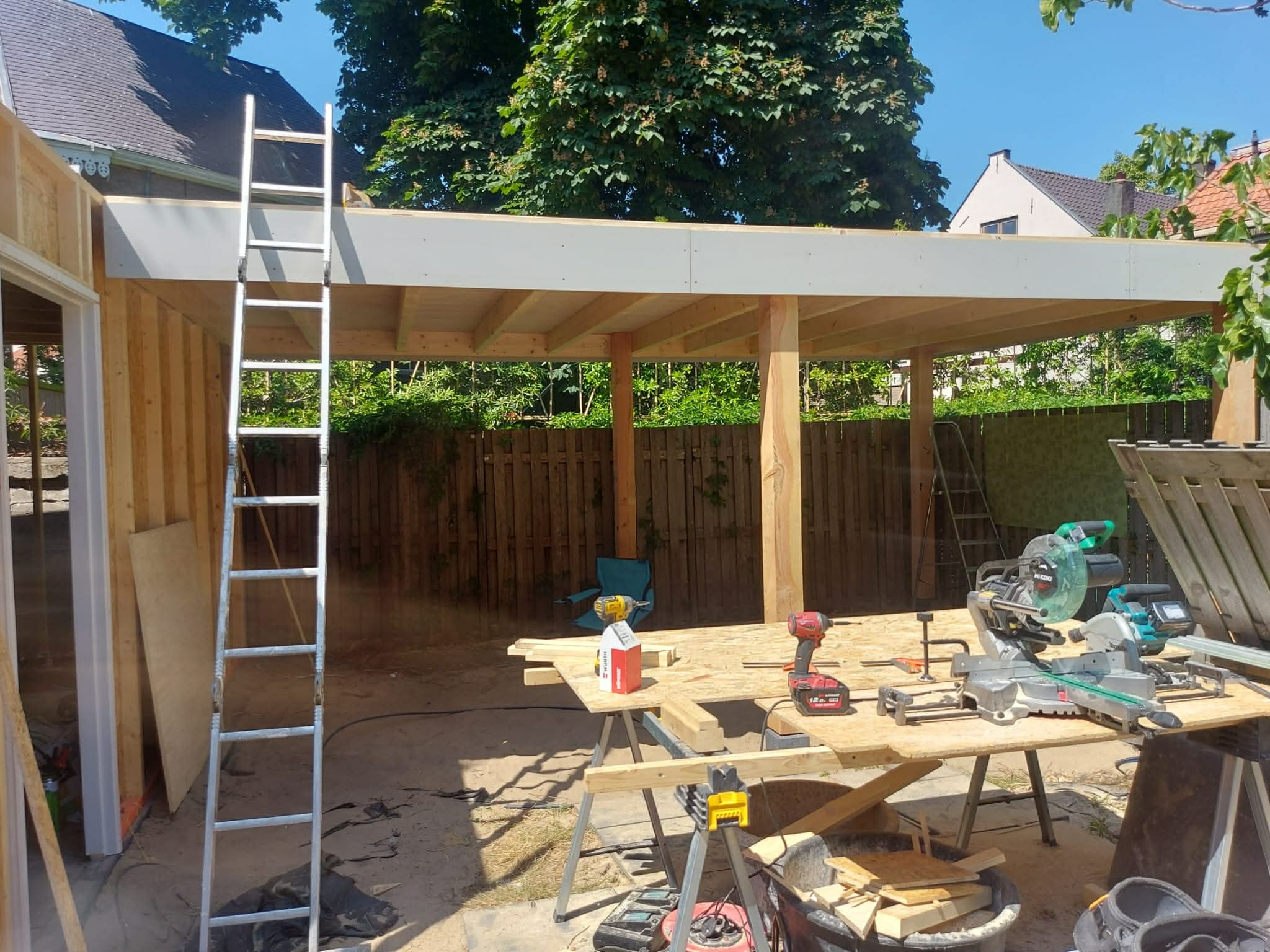 Konstrukcja drewniana z narzędziami w tle ogrodu. Widoczna drabina, piły i wkrętarki na stole roboczym. Elementy konstrukcyjne w trakcie montażu.