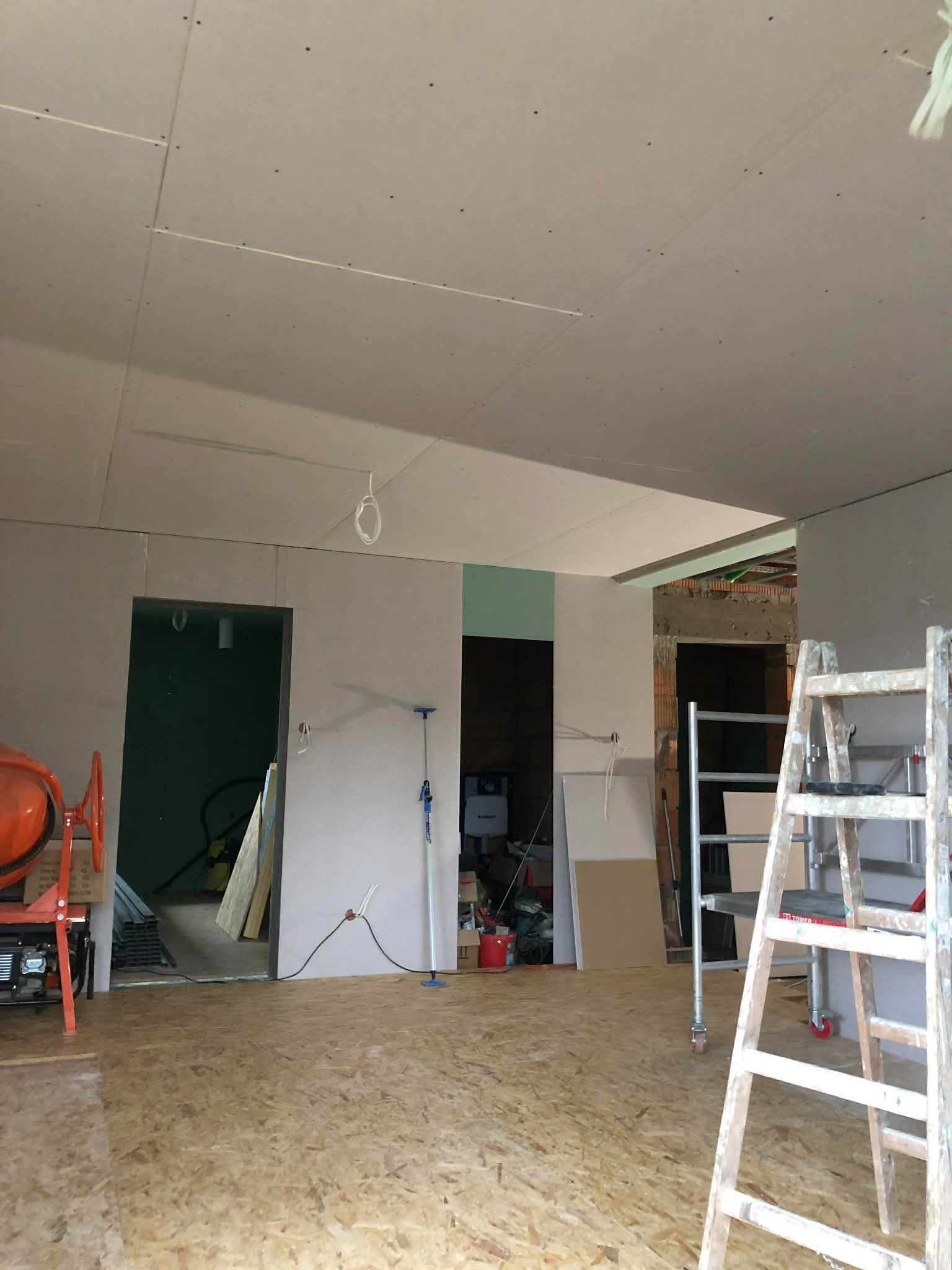 Wnętrze w trakcie prac wykończeniowych: zamontowane płyty gipsowo-kartonowe na suficie, widoczne narzędzia (mieszarka, drabina) i surowa podłoga z płyt OSB.