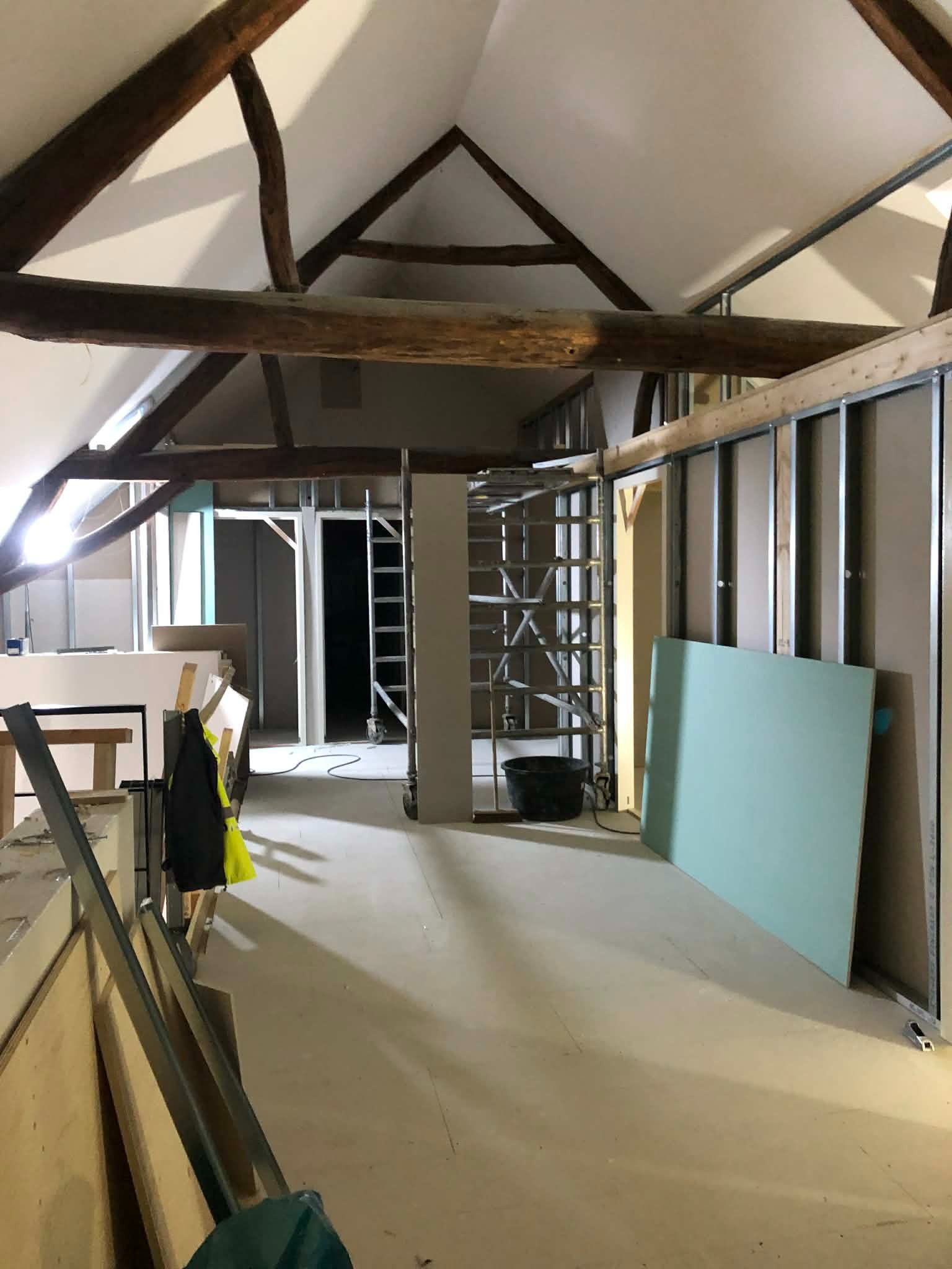 Poddasze w trakcie adaptacji: widoczne drewniane belki stropowe, metalowy stelaż pod płyty gipsowe i zielona płyta GK. W tle drabina i otwór drzwiowy.