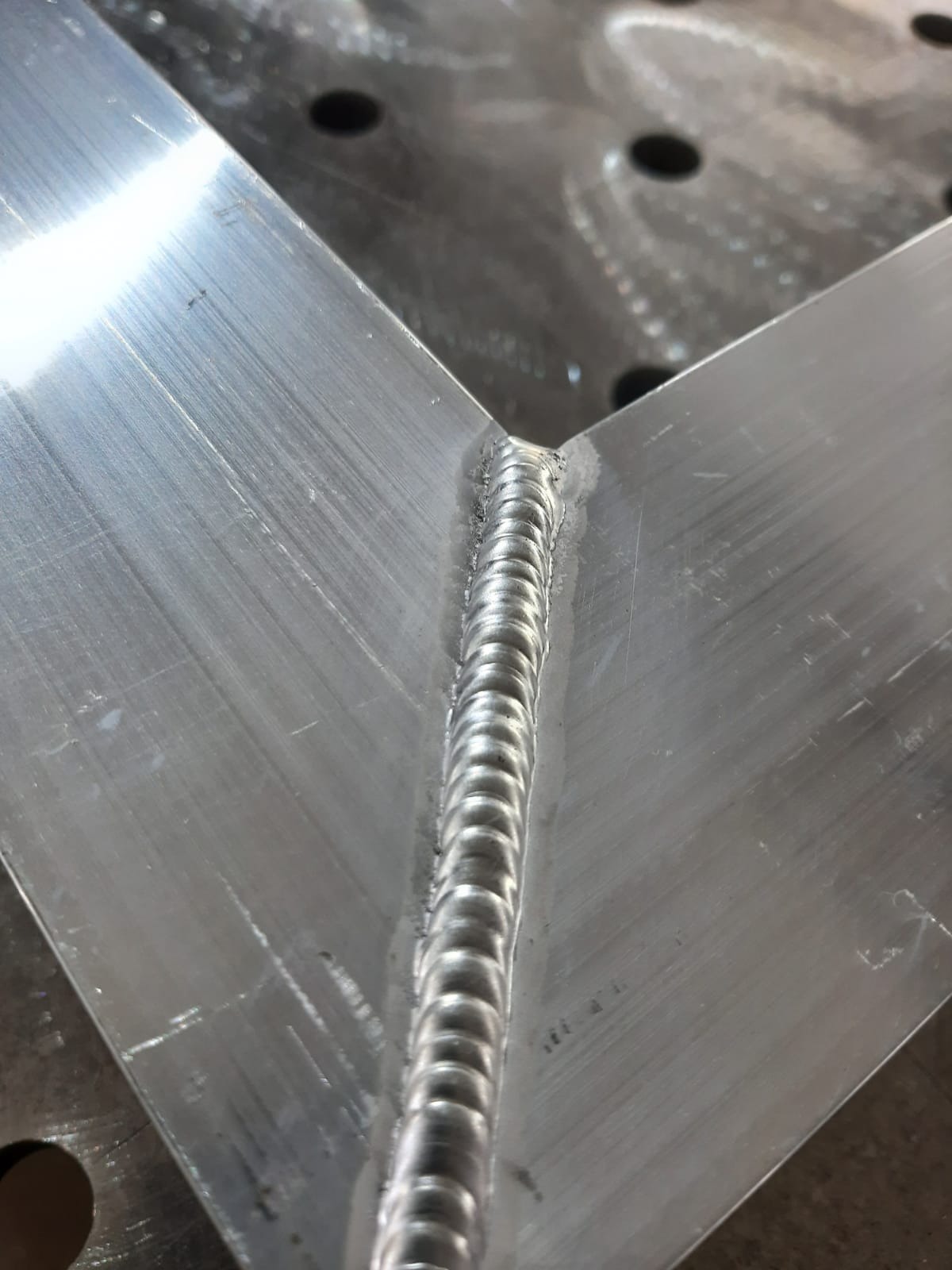 Połączenie spawane dwóch elementów aluminiowych pod kątem prostym, widoczne regularne spoiny na szlifowanej powierzchni metalu.