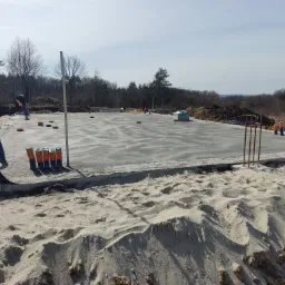 Wylana płyta betonowa na przygotowanym terenie budowy, widoczne wystające pręty zbrojeniowe, rury instalacyjne oraz robotnicy w oddali, drzewa w tle.