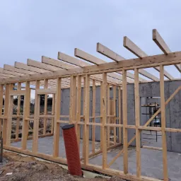 Surowy szkielet domu w trakcie budowy, widoczne drewniane belki, betonowe ściany fundamentowe i pomarańczowa rura drenażowa, pochmurne niebo w tle.