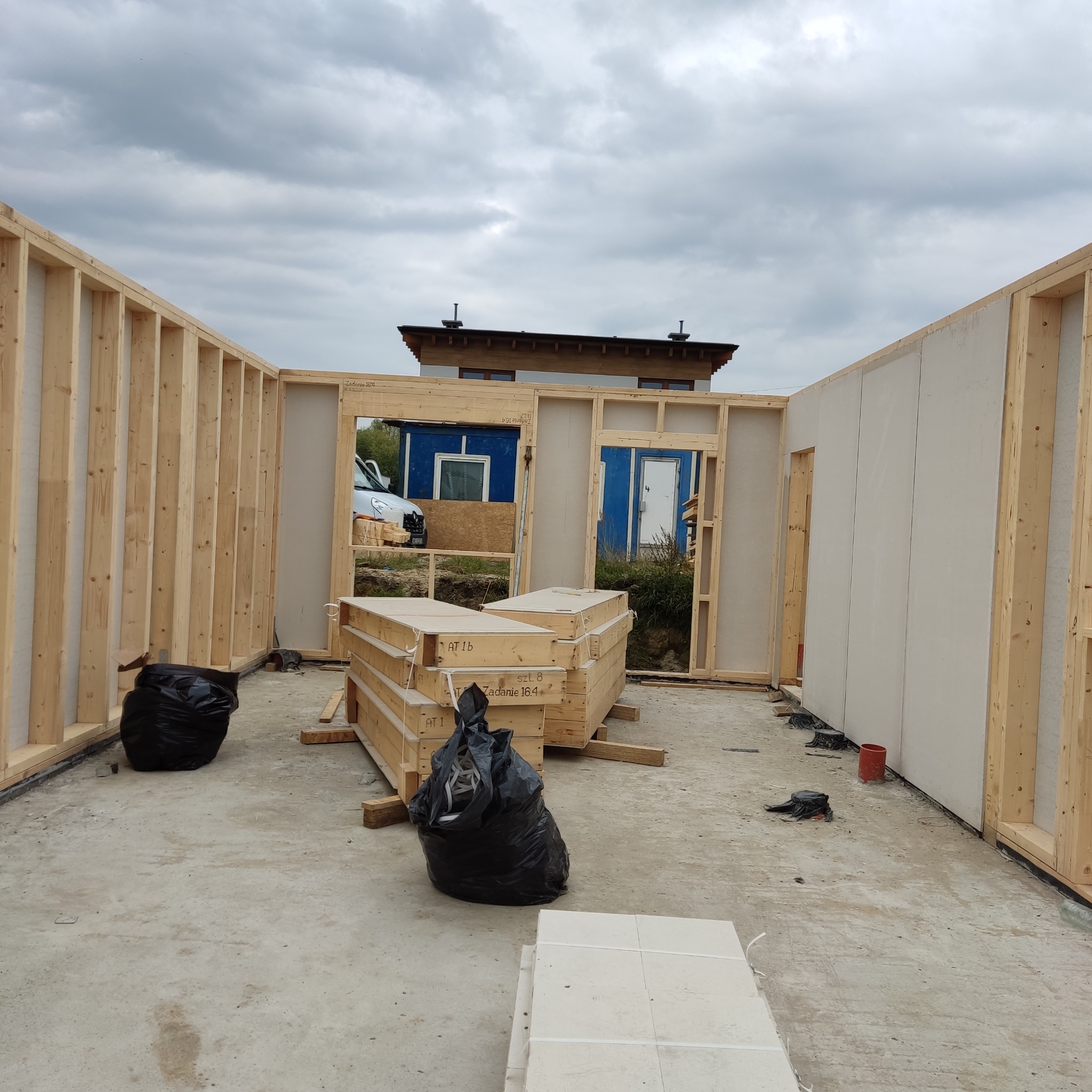 Wnętrze domu w trakcie budowy w technologii szkieletowej, widoczne drewniane ramy ścian, płyty OSB, czarne worki na śmieci i palety z materiałami budowlanymi, z zewnątrz widoczny budynek gospodarczy.