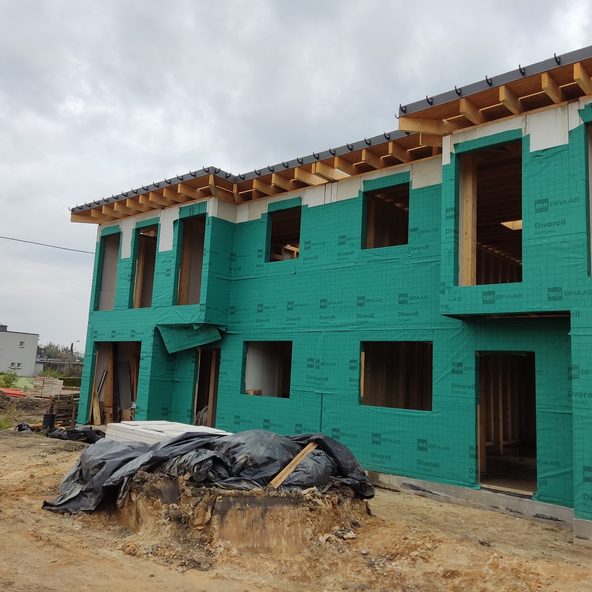 Wznoszony dwukondygnacyjny budynek szkieletowy z widoczną konstrukcją drewnianą, pokryty zieloną membraną paroizolacyjną z nadrukiem, widok z zewnątrz na placu budowy.