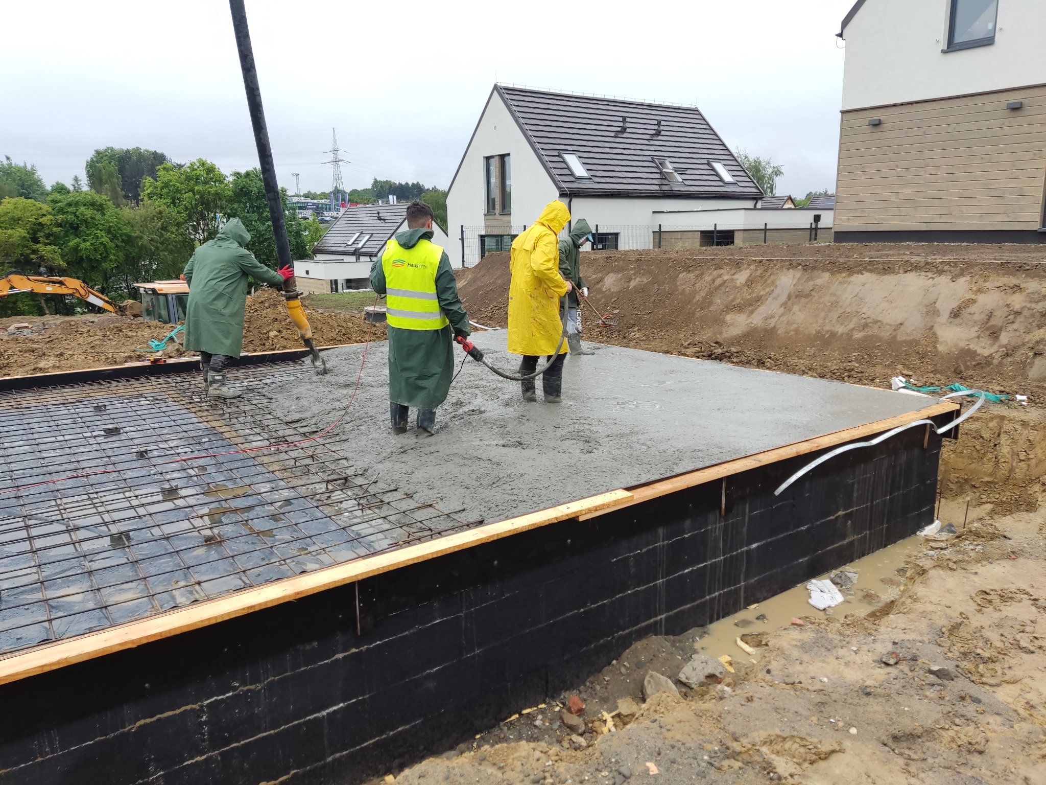 Wylewanie betonu na przygotowane zbrojenie fundamentu, pracownicy w odzieży ochronnej rozprowadzają beton z pompy, widoczne deskowanie i izolacja pionowa fundamentu.