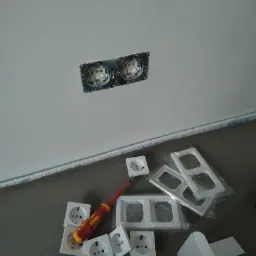 Montaż podwójnego gniazdka elektrycznego w ścianie, widoczne puszki instalacyjne, nowe gniazdka, ramki i izolowany śrubokręt na podłodze.
