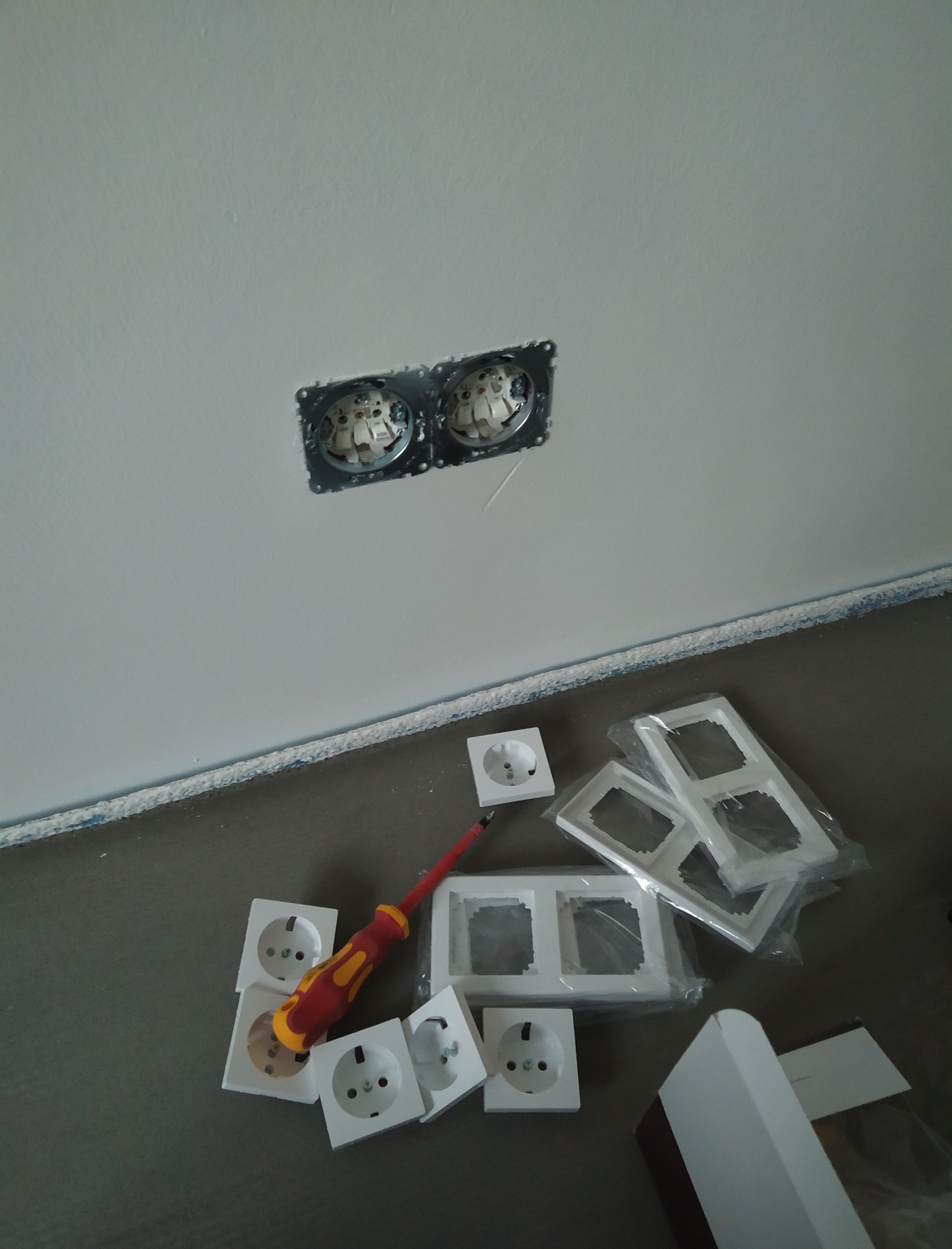 Montaż podwójnego gniazdka elektrycznego w ścianie, widoczne puszki instalacyjne, nowe gniazdka, ramki i izolowany śrubokręt na podłodze.