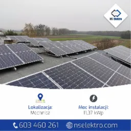 Instalacja paneli słonecznych na płaskim dachu w Nędzy. Widok z góry na liczne panele fotowoltaiczne z szarym niebem w tle. Na dalszym planie widoczne drzewa i pola.