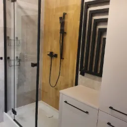 Nowoczesna kabina prysznicowa z czarnymi profilami i deszczownicą, drewnopodobna okładzina ścienna, grzejnik drabinkowy w stylu loft, jasne płytki i szafka łazienkowa.