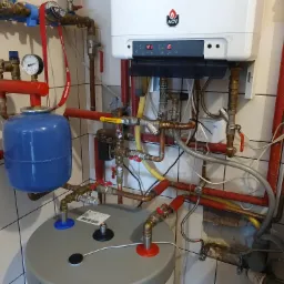 Kocioł grzewczy ACV z podłączonymi instalacjami, widoczny zbiornik wyrównawczy i zasobnik ciepłej wody w pomieszczeniu z kafelkami.