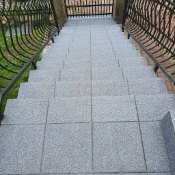 Zewnętrzne schody wyłożone szarymi płytkami granitowymi z czarną, kutą balustradą, prowadzące do bramy w Koszalinie.