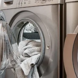 Srebrna pralka z otwartymi drzwiami, wewnątrz bęben wypełniony szarymi ręcznikami, widoczny panel sterowania u góry, obok druga pralka.