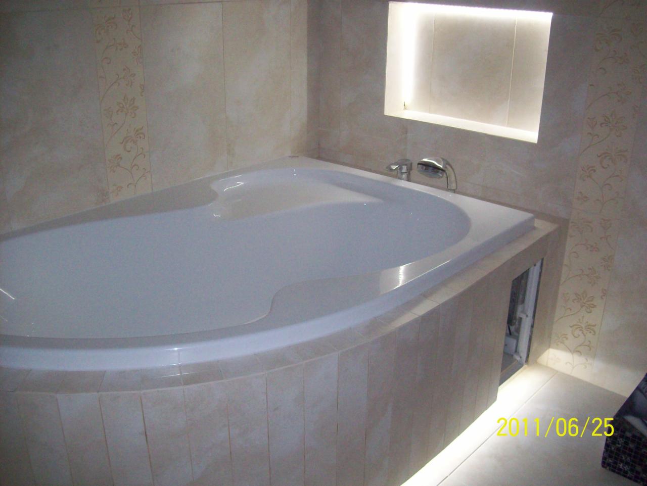 Wanna z hydromasażem obudowana beżowymi płytkami, z wnęką oświetloną od góry, armatura chromowana, beżowe płytki z delikatnym wzorem na ścianach.