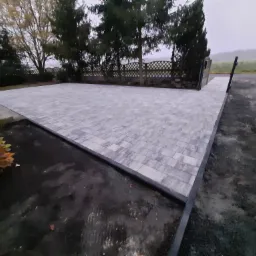 Ukończony podjazd z szarej kostki brukowej ułożonej we wzór jodełki, otoczony zielenią i ogrodzeniem panelowym.