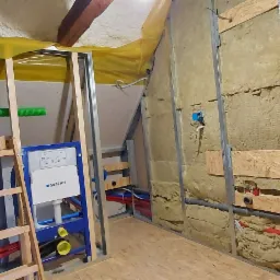 Montaż stelaża podtynkowego Geberit w łazience na poddaszu, widoczne izolacja ścian wełną mineralną, rury instalacyjne i fragment konstrukcji sufitu.