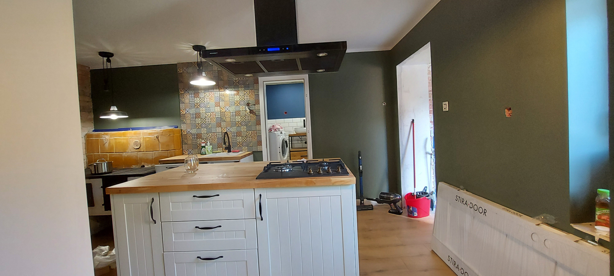 Wnętrze kuchni w trakcie remontu: widoczna wyspa kuchenna z drewnianym blatem i białymi szafkami, stara kuchnia kaflowa w tle, ściana z dekoracyjnymi płytkami oraz przygotowane do montażu nowe drzwi.