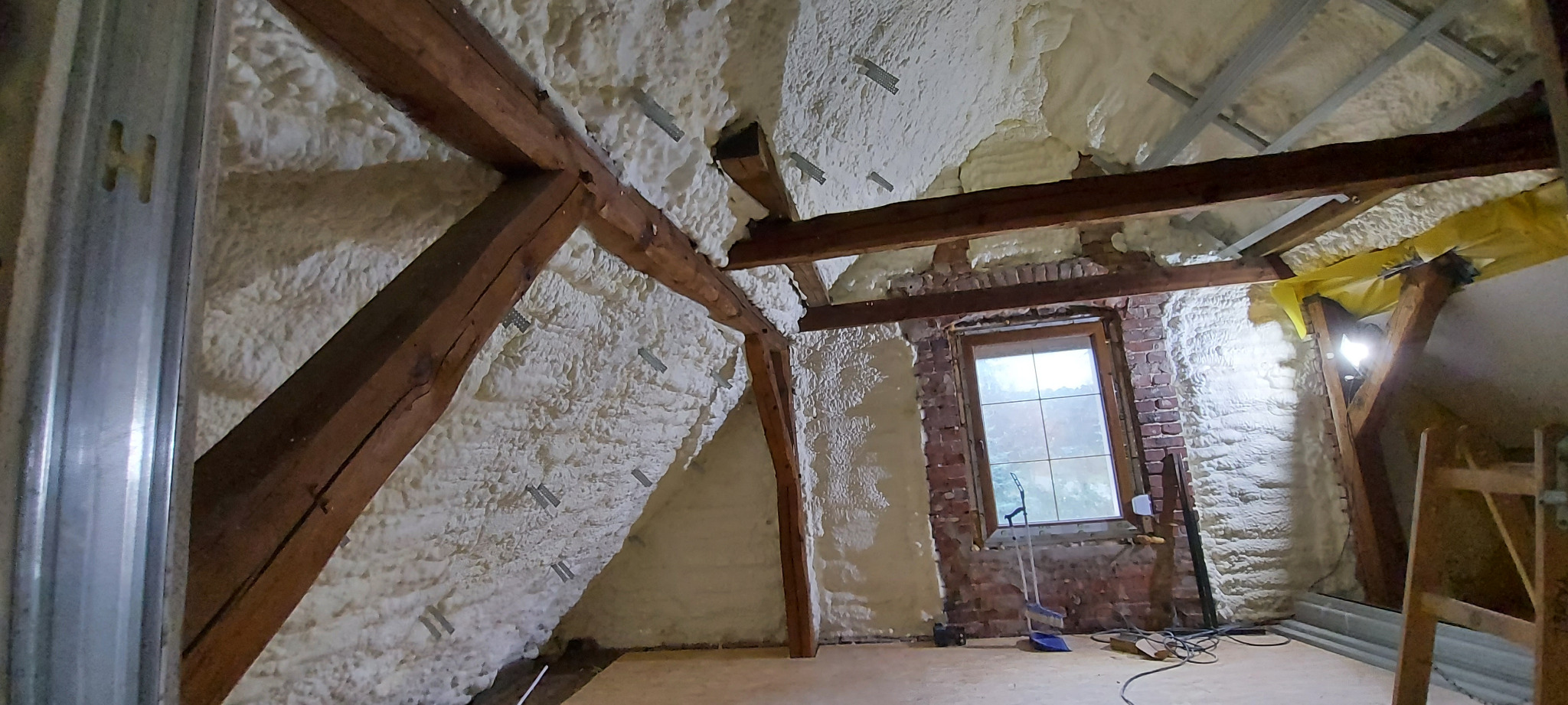 Poddasze w trakcie ocieplania pianą poliuretanową, widoczne drewniane belki stropowe, fragment ściany z cegły z oknem oraz elementy konstrukcji metalowej.