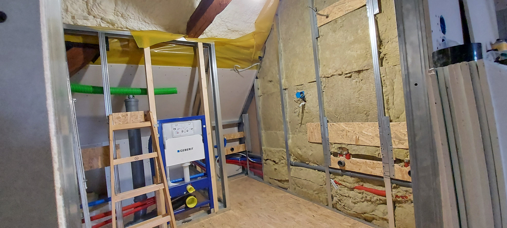 Montaż stelaża podtynkowego Geberit w łazience na poddaszu, widoczne izolacja ścian wełną mineralną, rury instalacyjne i fragment konstrukcji sufitu.