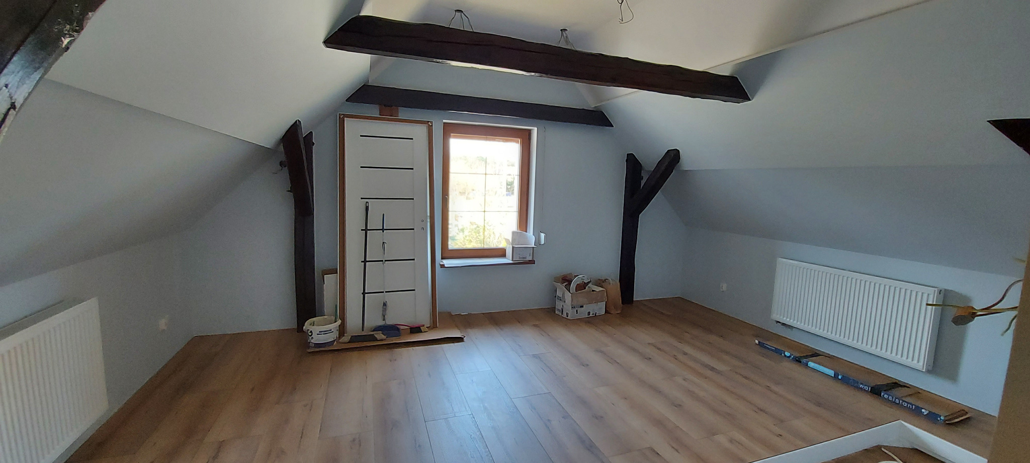 Poddasze po remoncie: nowa podłoga laminowana, świeżo pomalowane ściany na szaro, białe grzejniki, drewniane belki stropowe i przygotowana do montażu biała ościeżnica z drzwiami.