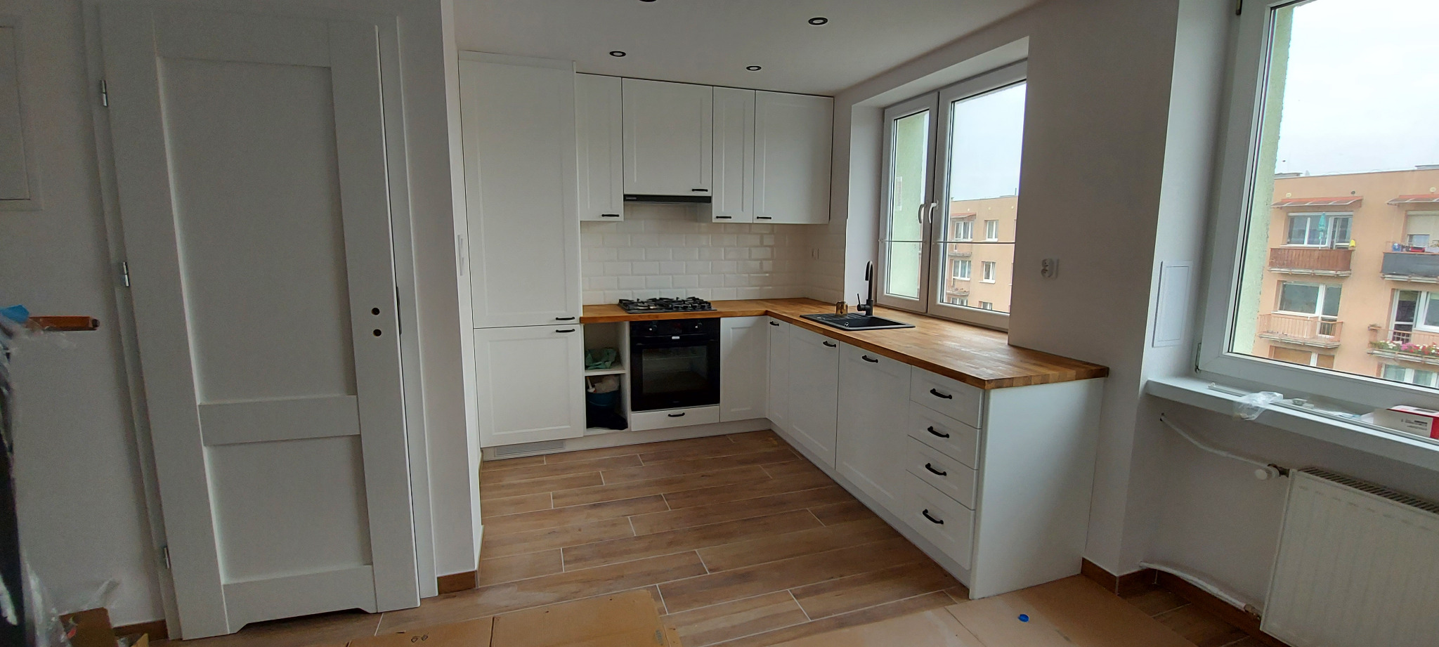 Nowoczesna, jasna kuchnia w stylu skandynawskim z białymi szafkami, drewnianym blatem i płytkami metro, widok na budynek mieszkalny za oknem.