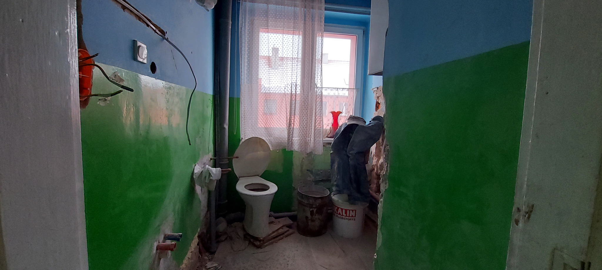 Łazienka w trakcie remontu: ściany pomalowane do połowy na zielono i niebiesko, odsłonięte kable elektryczne, rury, toaleta i wiadra z farbą w pomieszczeniu z oknem z firanką.