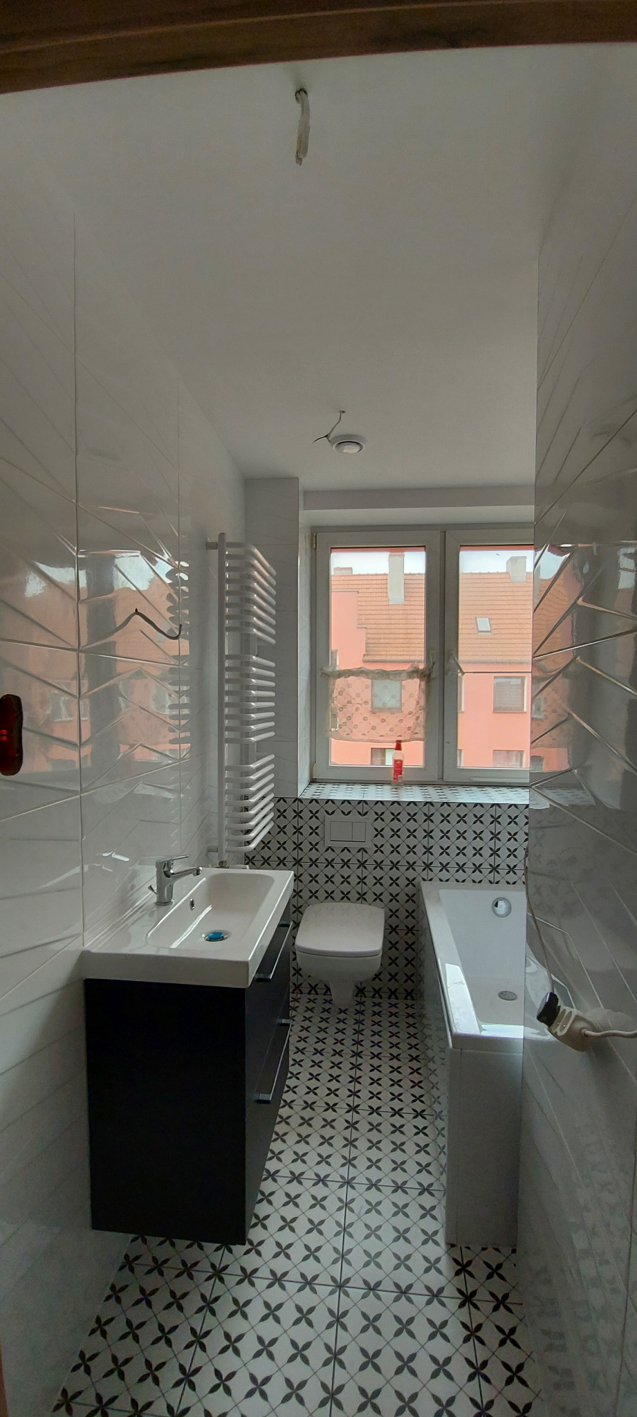 Wykończona łazienka z biało-czarną mozaiką na podłodze i ścianie w strefie toalety i wanny, białe płytki z fakturą na pozostałych ścianach, czarna szafka pod umywalką i grzejnik drabinkowy.