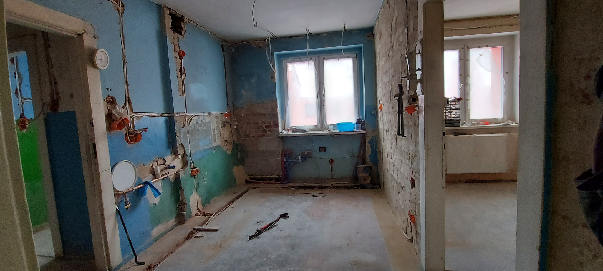 Wnętrze mieszkania w trakcie remontu: odsłonięte instalacje elektryczne i hydrauliczne, częściowo skuwa ściana, widoczne przewody i puszki elektryczne, niebieskie ściany z resztkami farby...
