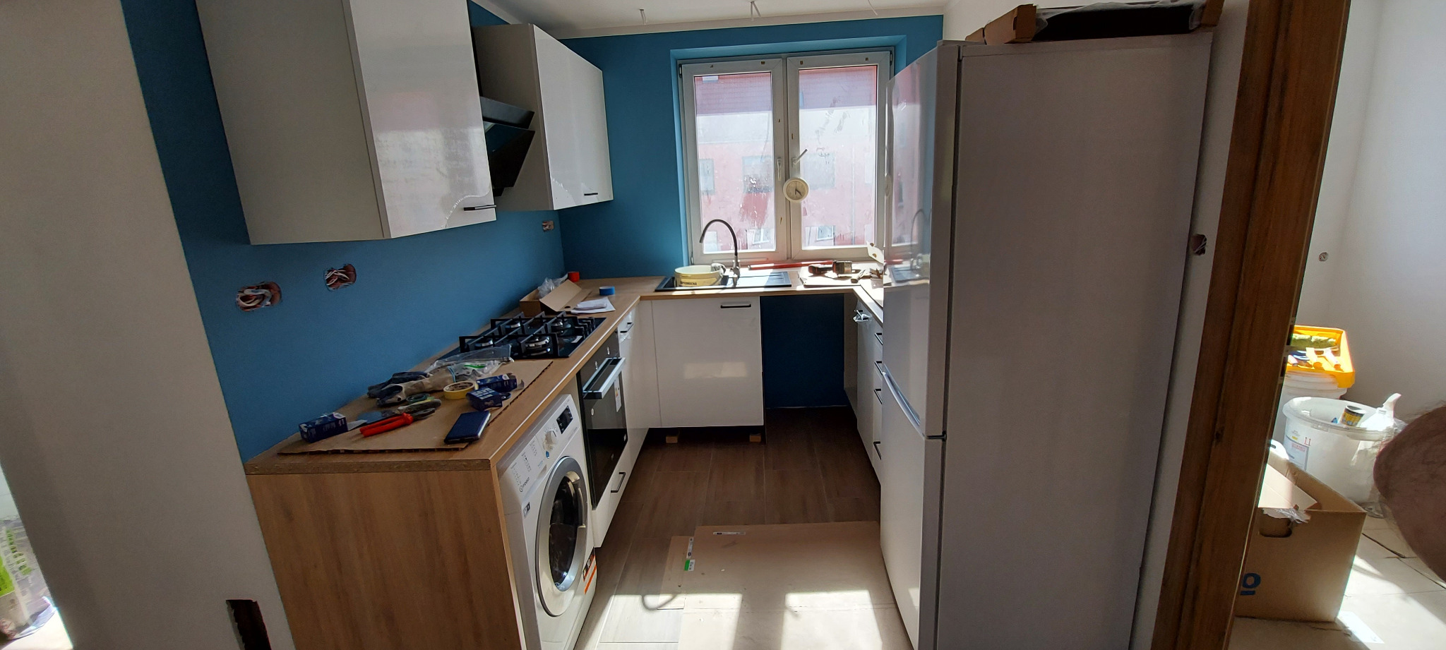 Wnętrze kuchni w trakcie remontu: niebieska ściana, białe szafki, sprzęt AGD częściowo zamontowany, widoczne narzędzia i kable przy gniazdkach.