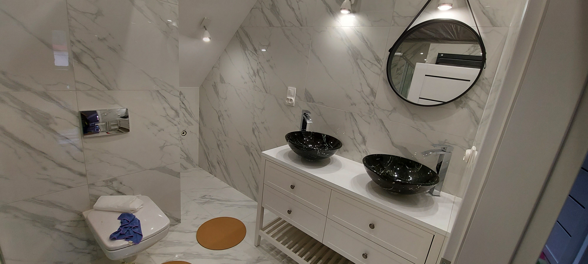 Wykończona łazienka na poddaszu z marmurowymi płytkami, dwiema czarnymi umywalkami nablatowymi, okrągłym lustrem w czarnej ramie i brązowym dywanikiem na podłodze.