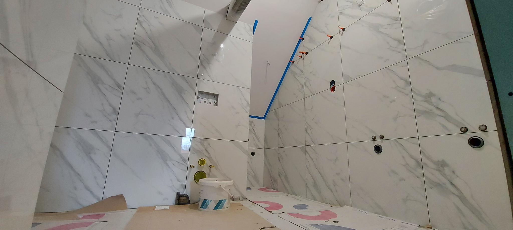 Wnętrze łazienki w trakcie remontu: białe, marmurowe płytki ścienne, widoczne otwory na instalacje, poziomowanie płytek, zabezpieczona podłoga. Wiadro z klejem i poziomica na podłodze.