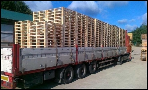 Ciężarówka z pełną naczepą drewnianych palet, widok z boku, na placu składowym w słoneczny dzień.