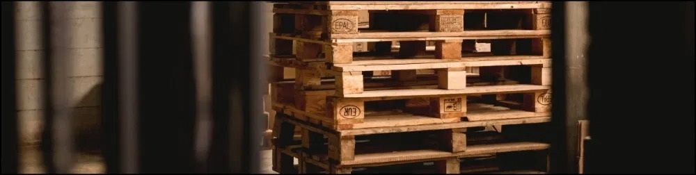 Stos drewnianych palet EUR w magazynie, widoczne oznaczenia licencyjne EPAL, ujęcie zza częściowo zasłaniającej konstrukcję.