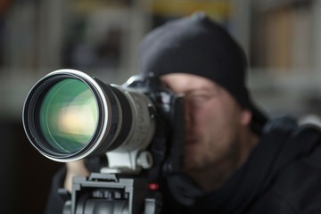 Mężczyzna w czapce robiący zdjęcie profesjonalnym aparatem z dużym obiektywem na statywie, z widocznym odbiciem w szkle obiektywu.