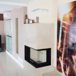 Nowoczesny, narożny wkład kominkowy z obudową z jasnego kamienia, czarna ramka, szklane drzwiczki, błyszcząca podłoga, dekoracje na górze, abstrakcyjna grafika na ścianie.