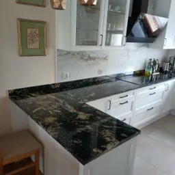 Czarny blat kamienny z wyraźnym złotym użyleniem w nowoczesnej białej kuchni z szafkami z przeszklonymi frontami i marmurowym backsplashiem, widoczne geometryczne lampy wiszące i drewniany stołek...