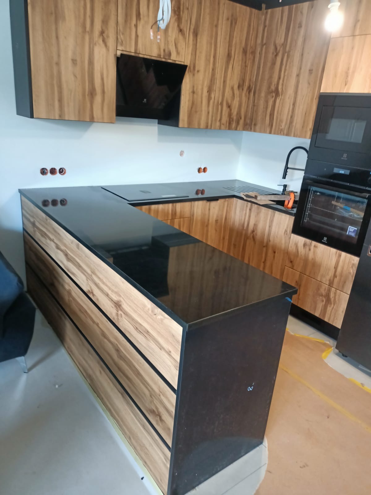 Czarny, błyszczący blat w kuchni z drewnianymi frontami szafek i widocznymi gniazdkami elektrycznymi w ścianie, jeszcze przed ostatecznym wykończeniem.