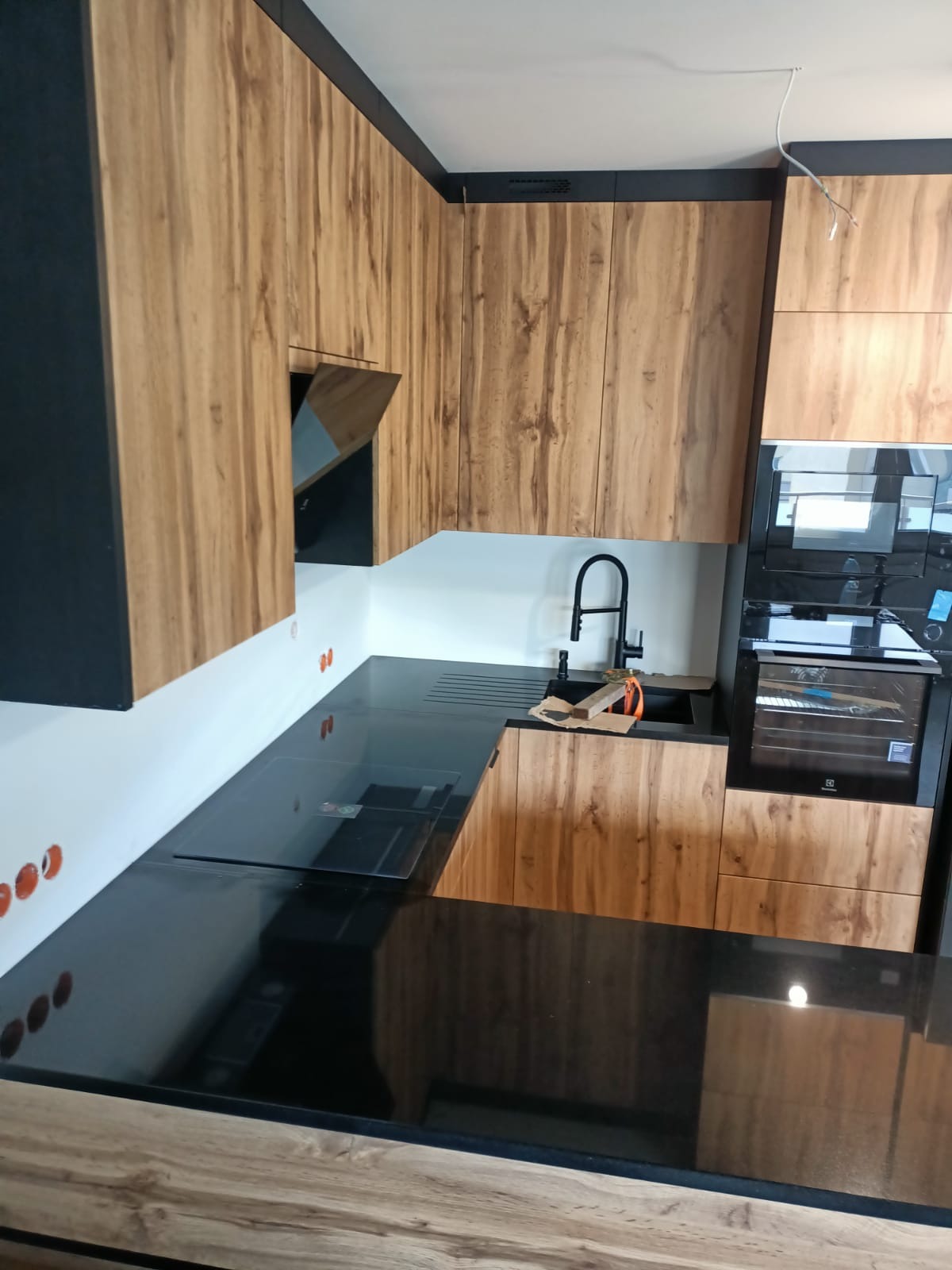 Nowoczesna kuchnia z czarnym blatem i drewnopodobnymi szafkami, czarny zlew i bateria, widoczne gniazdka elektryczne na ścianie.