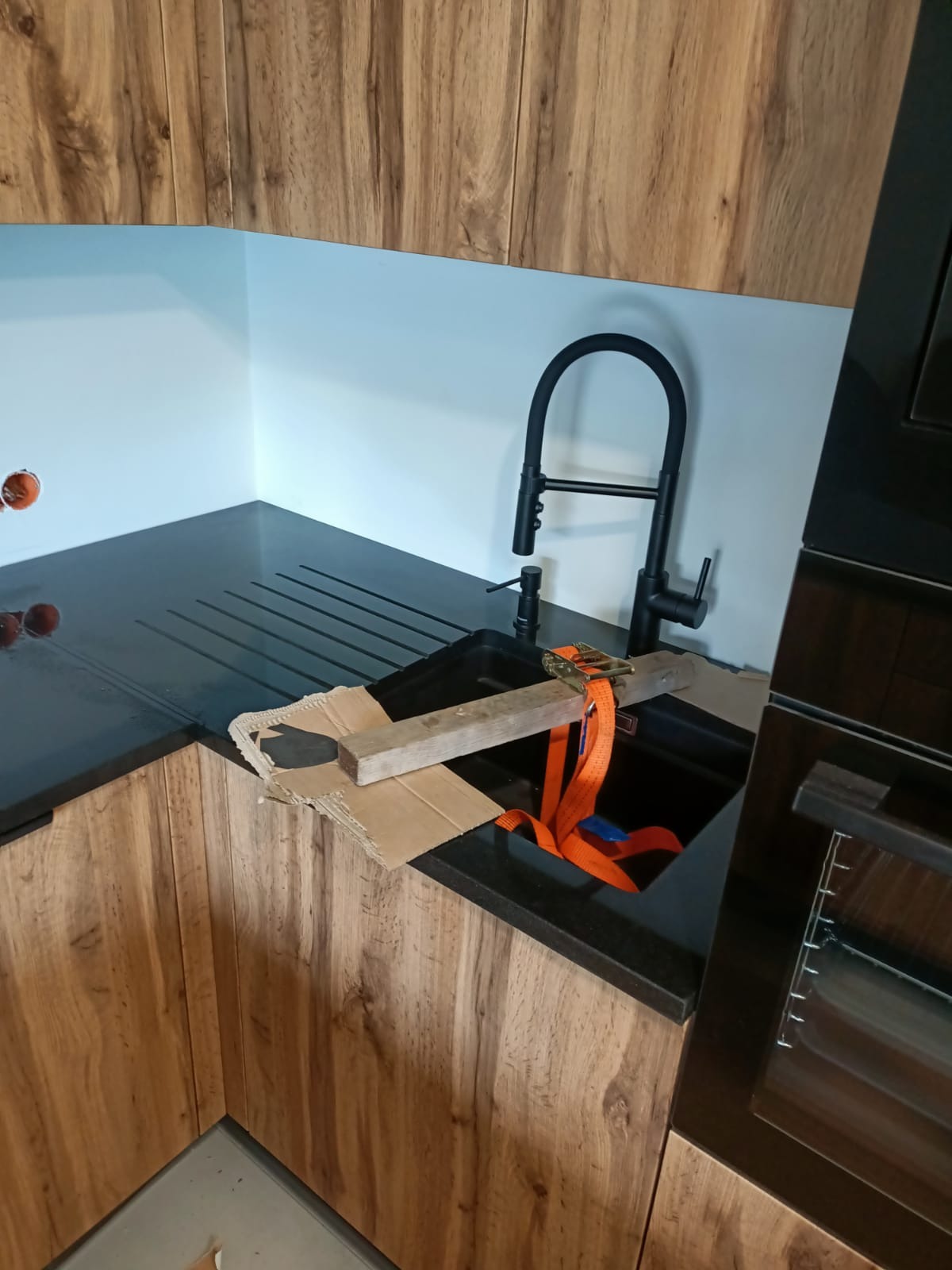 Montaż czarnego zlewu w blacie kuchennym z ciemnego kamienia, zabezpieczonego deską i pasem transportowym w trakcie instalacji.