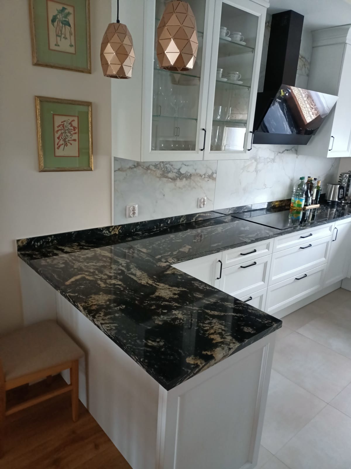 Czarny blat kamienny z wyraźnym złotym użyleniem w nowoczesnej białej kuchni z szafkami z przeszklonymi frontami i marmurowym backsplashiem, widoczne geometryczne lampy wiszące i drewniany stołek...