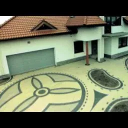 Widok z góry na dom z jasną elewacją i garażem, otoczony ozdobnym chodnikiem z kostki brukowej ułożonej we wzory geometryczne, w tym duży okrąg z motywem roślinnym.