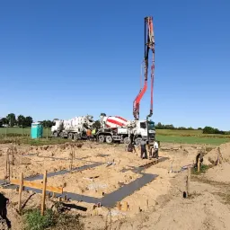 Wylewanie betonu do szalunków fundamentowych pod budowę domu jednorodzinnego, widoczne betoniarki i pompa do betonu na tle pola uprawnego.