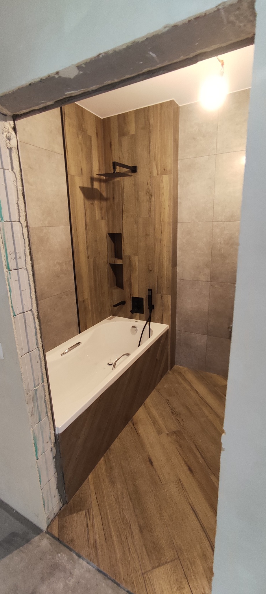 Nowoczesna łazienka w trakcie remontu: biała wanna z czarną armaturą, imitacja drewna na ścianach i podłodze, widoczny surowy fragment ściany przy wejściu.