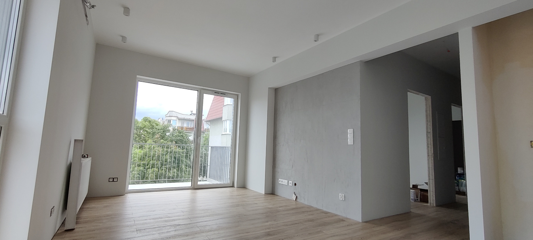 Wnętrze jasnego pokoju z dużym oknem balkonowym, widok na zieleń za oknem, panele podłogowe, ściany w odcieniach szarości, widoczne gniazdka elektryczne i włączniki.