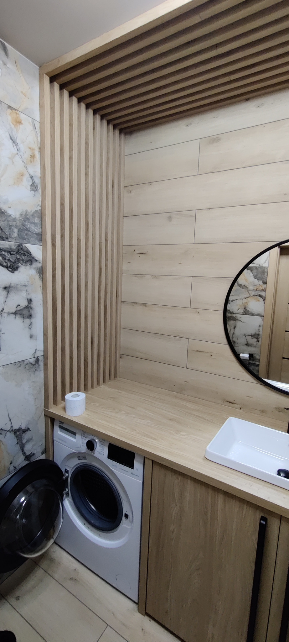 Nowoczesna łazienka z drewnianymi lamelami na suficie i ścianie, marmurową płytką, drewnianym blatem z umywalką, pralką z otwartymi drzwiami i okrągłym lustrem.
