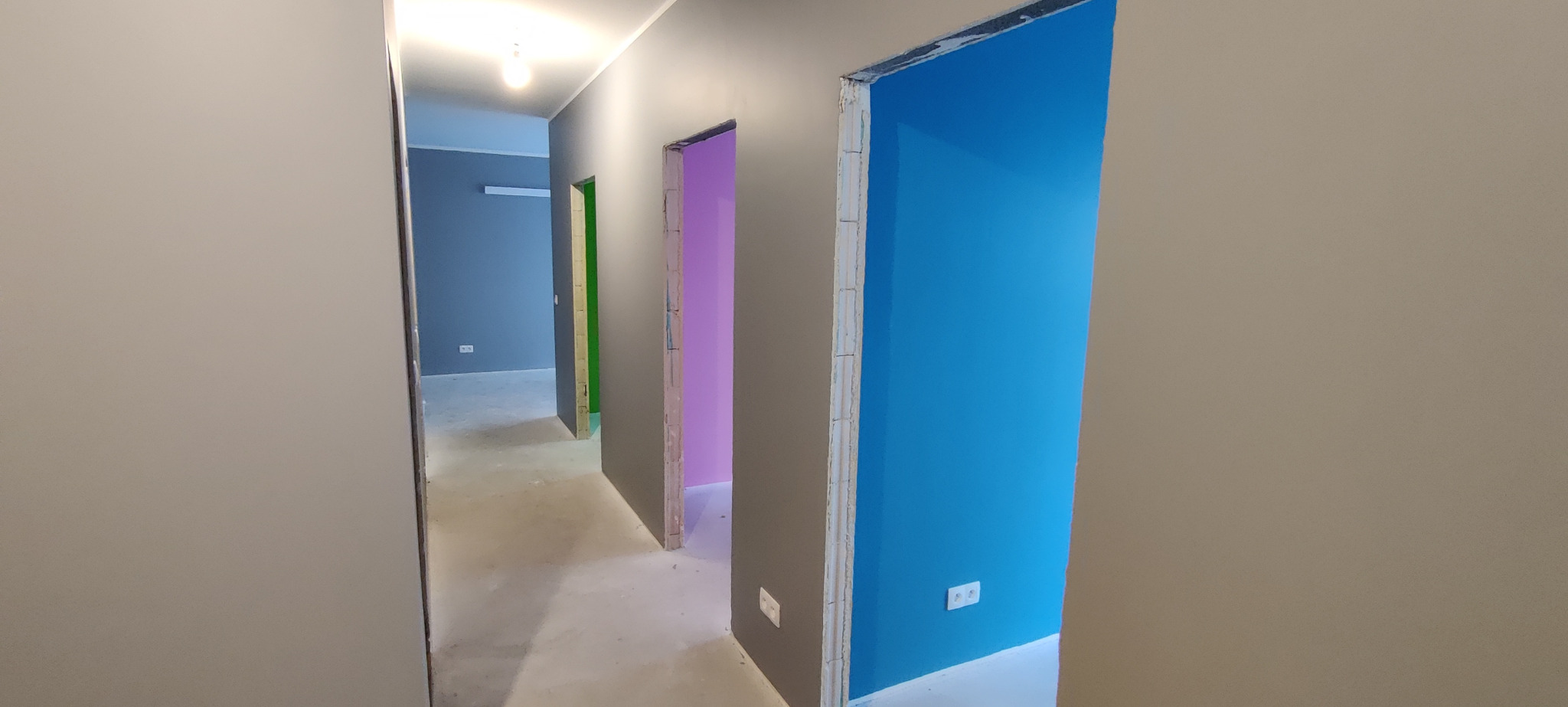 Wnętrze mieszkania w Bydgoszczy w trakcie malowania, widoczne ściany w różnych kolorach (niebieski, różowy, zielony), szare futryny drzwiowe, surowa podłoga i gniazdka elektryczne.