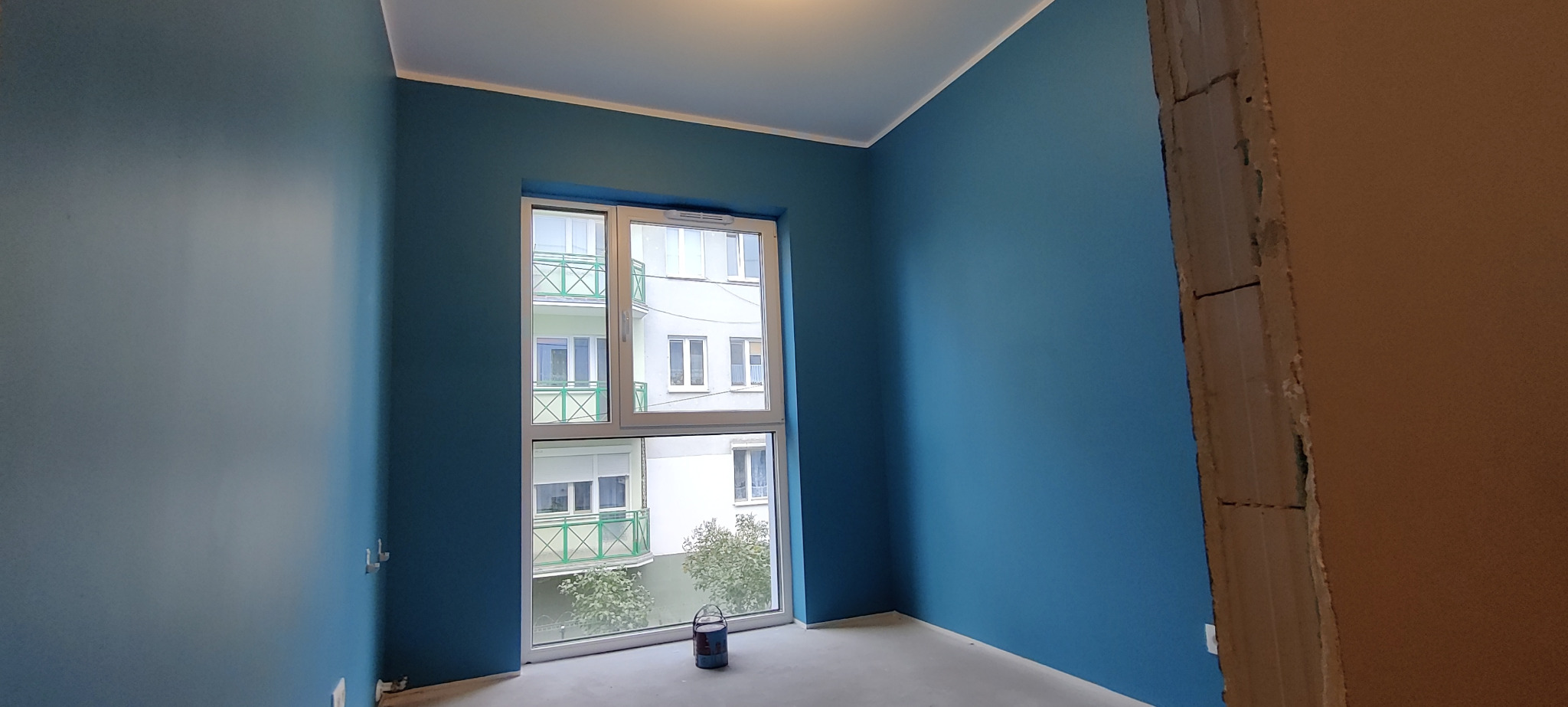 Pomieszczenie po malowaniu ścian na kolor morski z widokiem na blok mieszkalny przez okno, puszka farby stoi na podłodze, widoczne surowe wykończenie futryny.