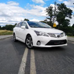 Biała Toyota Avensis kombi zaparkowana na asfalcie z podwójną linią, z widocznym znakiem drogowym i drzewami w tle pod błękitnym niebem z chmurami.